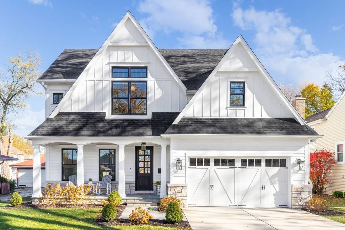 A white modern farmhouse with a dark shingled roof and black window frames. The bottom of the house has a light rock siding and covered front porch